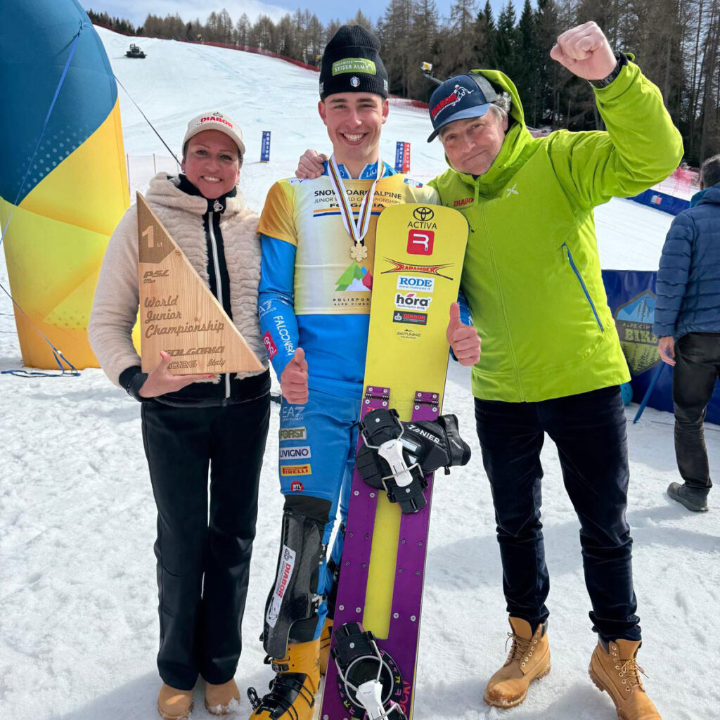 Tre persone su una pista da snowboard: al centro un giovane atleta con medaglia e tavola, alla sua sinistra una donna con un trofeo a forma di triangolo che riporta “1st – World Junior Championship – Folgaria 2025 Italy”, alla destra un uomo in giacca verde che alza il pugno in segno di vittoria.