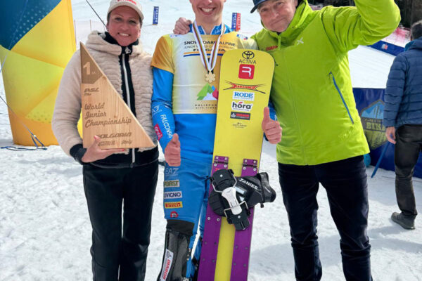 Tre persone su una pista da snowboard: al centro un giovane atleta con medaglia e tavola, alla sua sinistra una donna con un trofeo a forma di triangolo che riporta “1st – World Junior Championship – Folgaria 2025 Italy”, alla destra un uomo in giacca verde che alza il pugno in segno di vittoria.